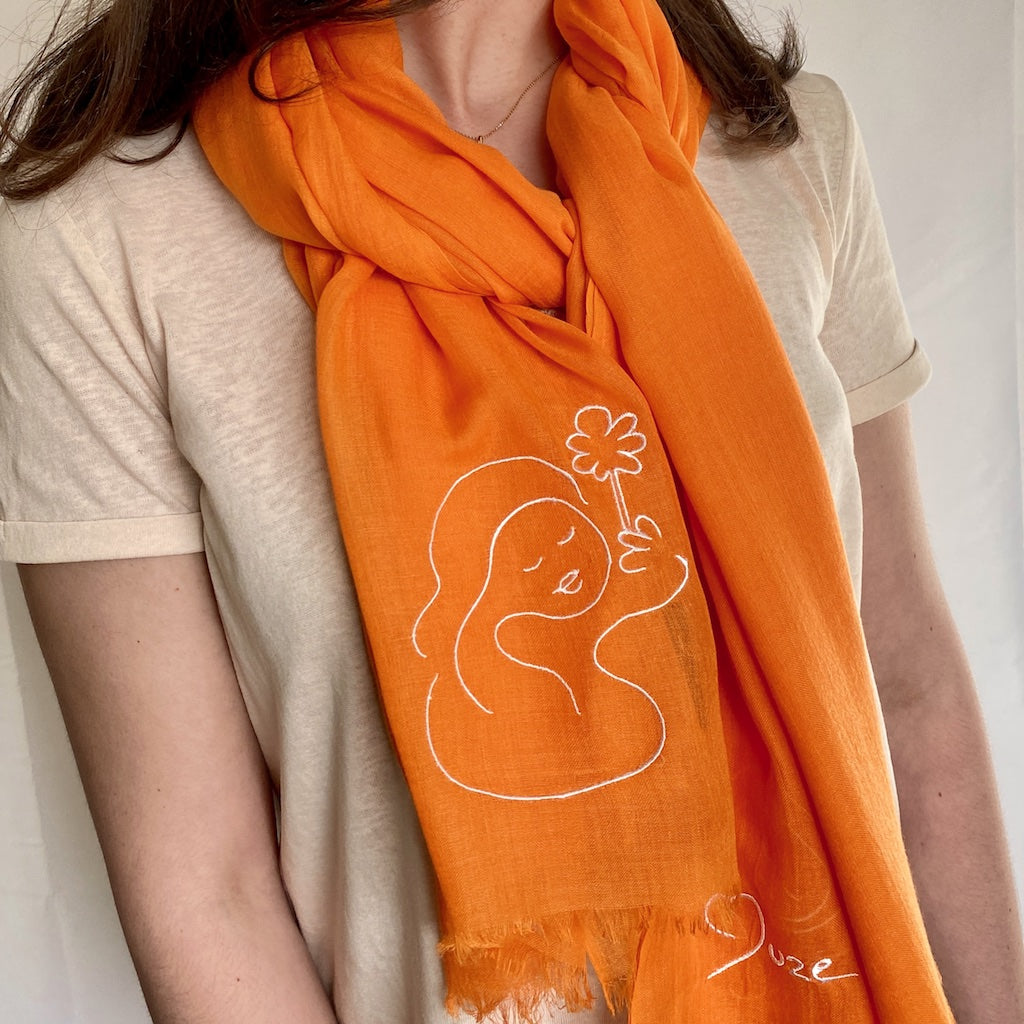 Muze orange lightweight scarf in modal and silk on model displaying embroidery of muze peace original artistic drawing on one side and Muze logo on other